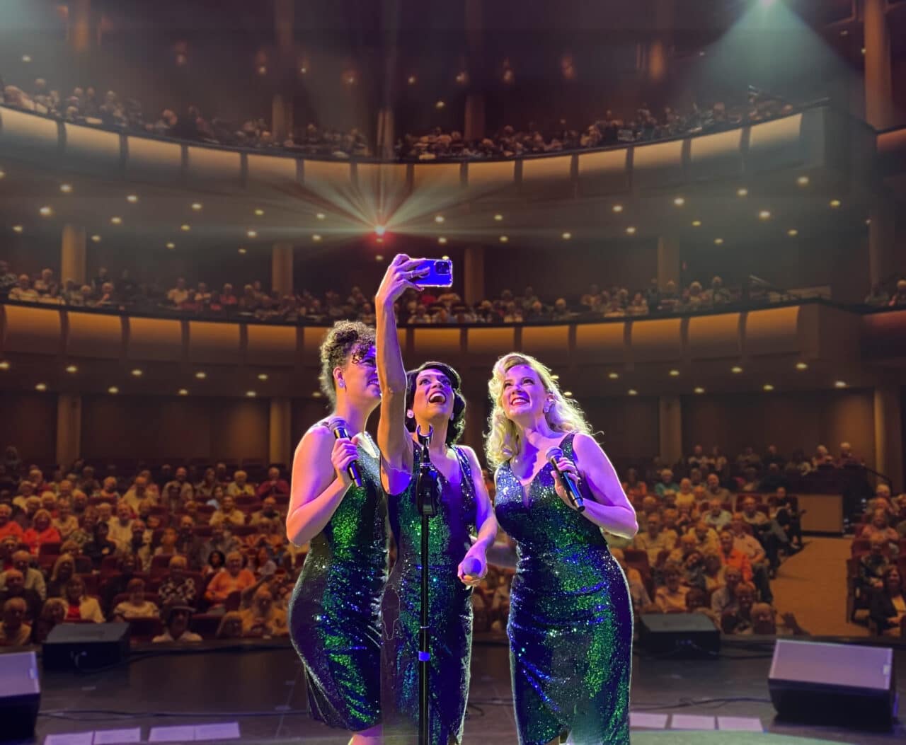 The Starlets on stage taking a selfie with a full audience in a performing arts center